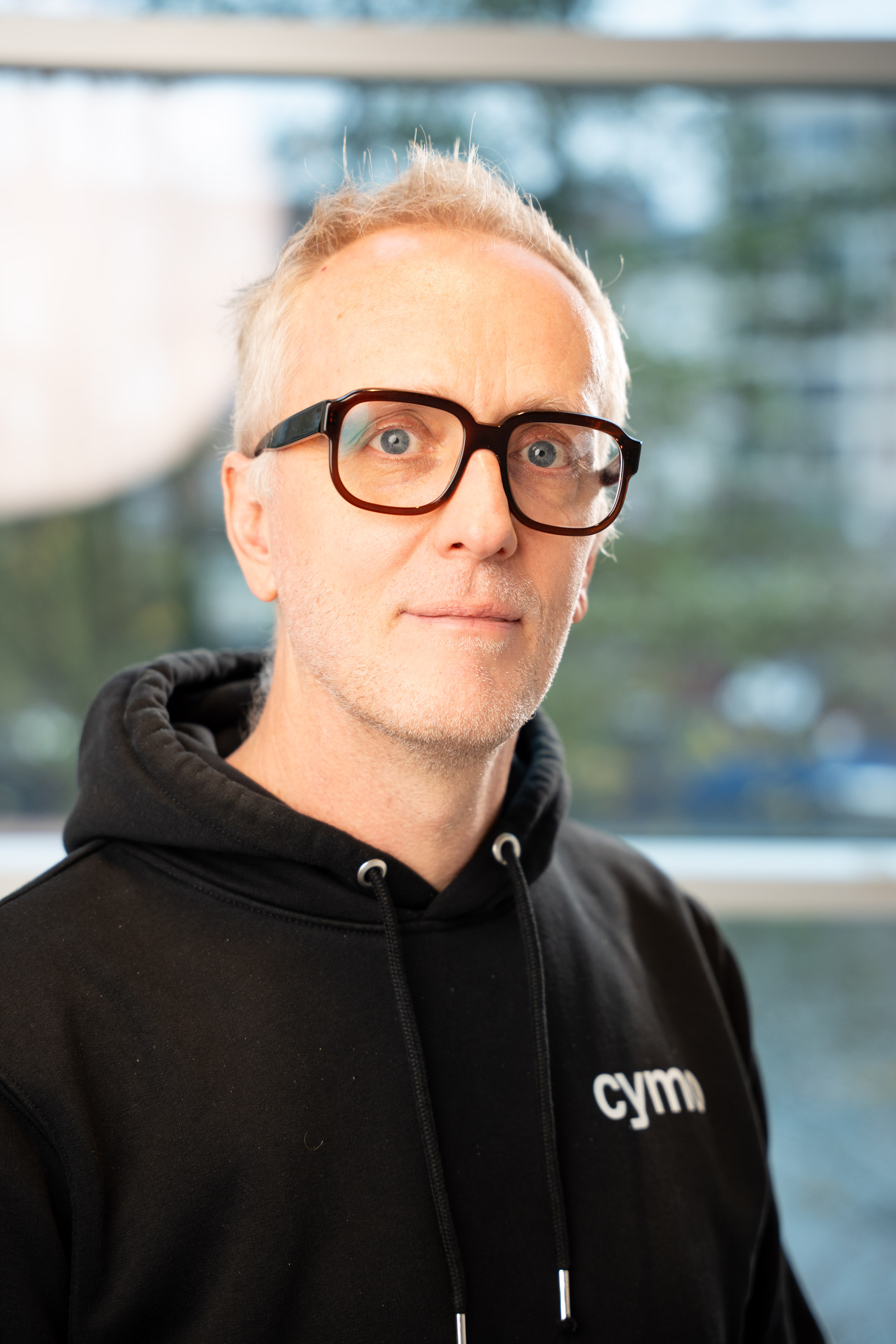 Man with short gray hair and glasses wearing a black hoodie with 'cym' logo, standing indoors near window.