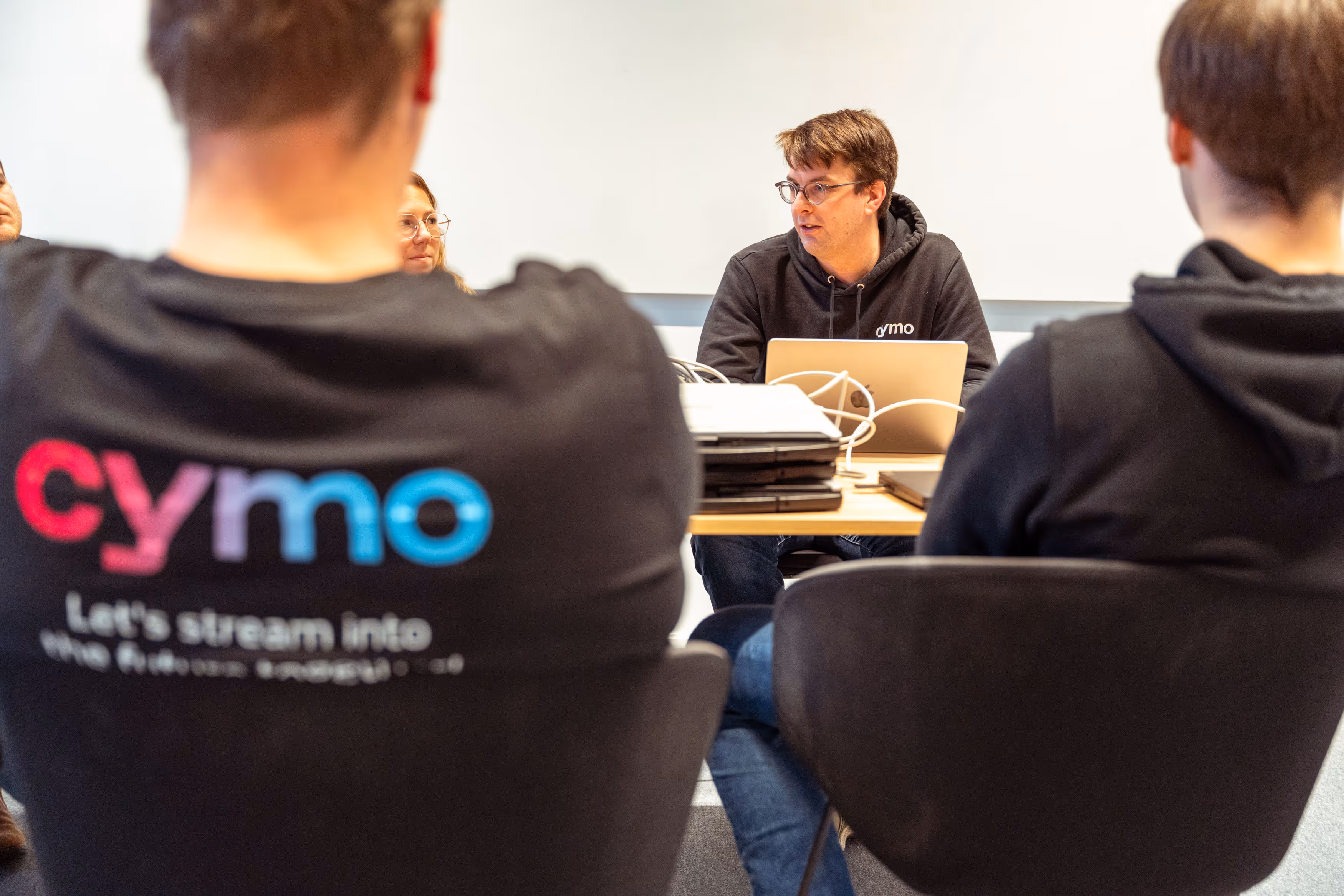 A man wearing glasses and a black Cymo hoodie speaking at a table with laptops, viewed from behind two other people also wearing black hoodies.