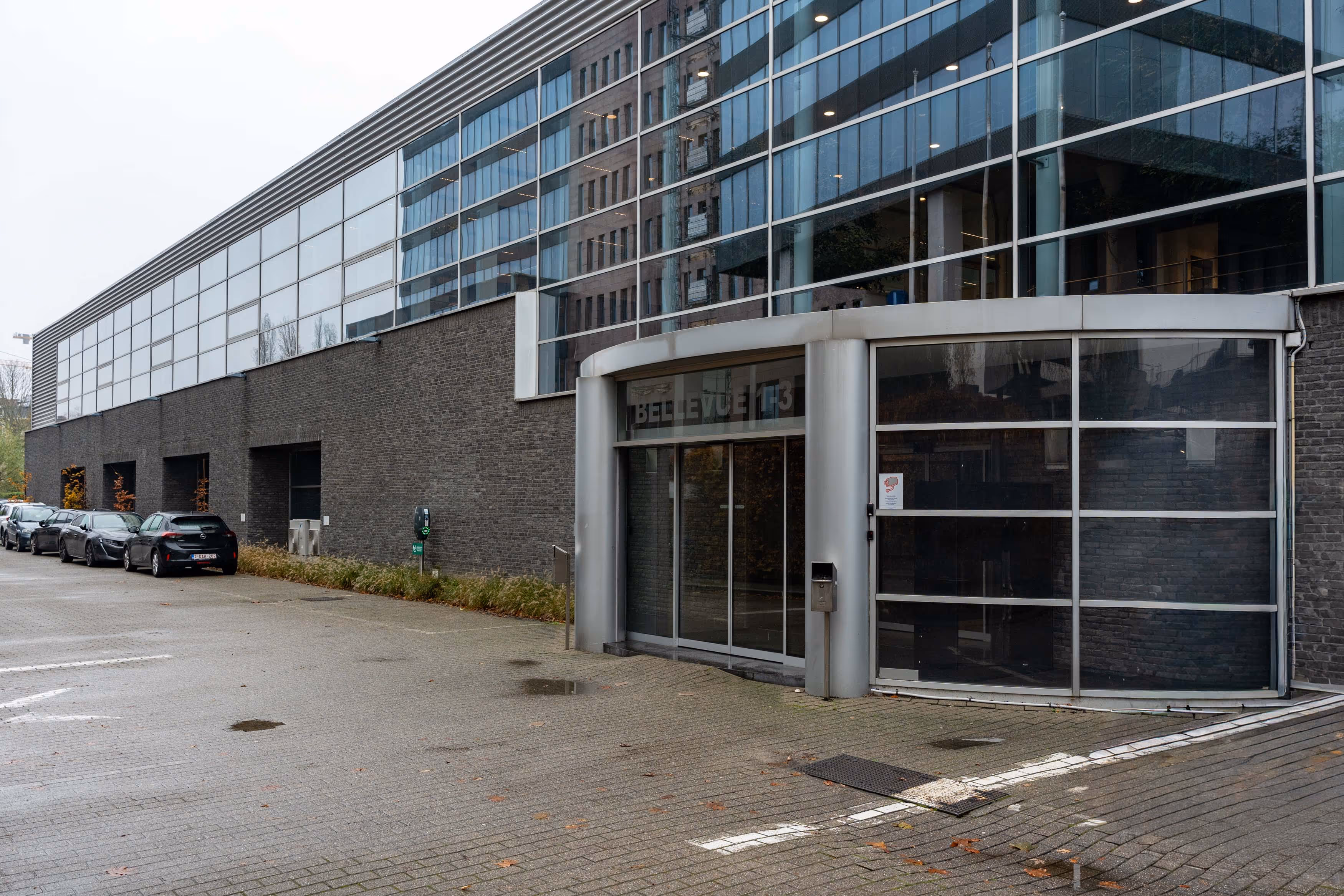 Modern office building entrance with glass and metal facade, cars parked along the side.