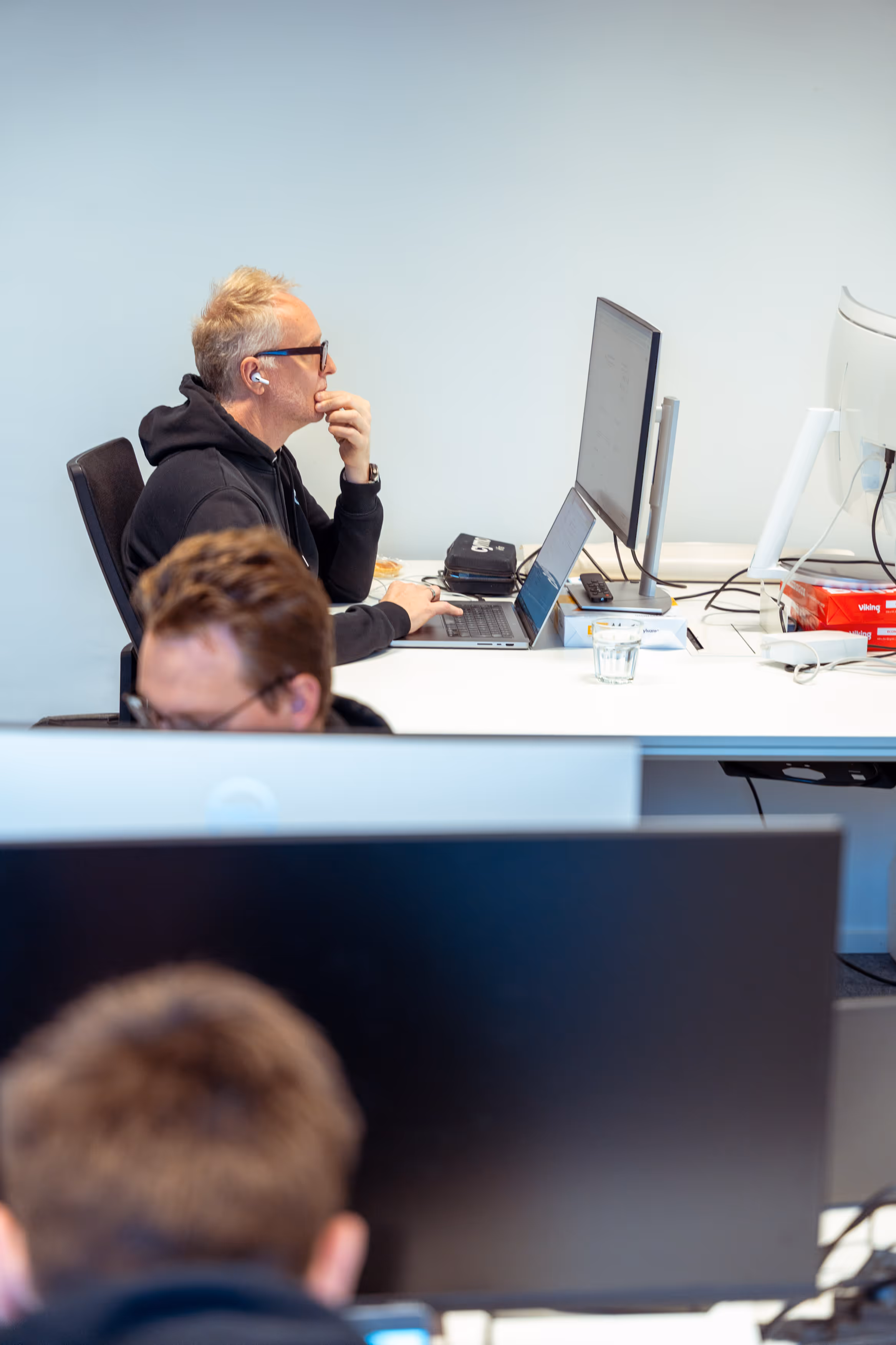 Man wearing glasses and earbuds working on a laptop at a white office desk with multiple monitors and office supplies.