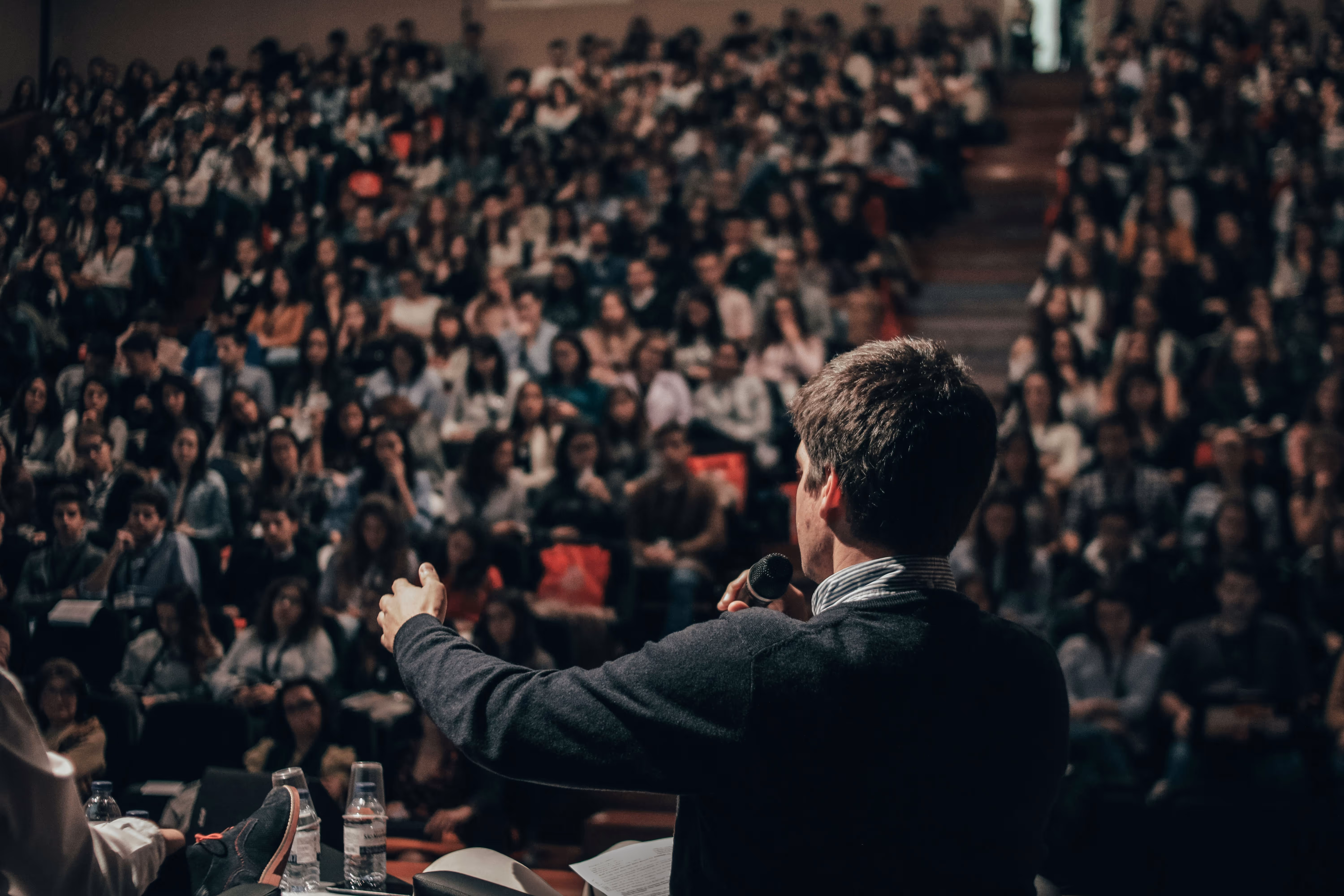 Man speaking into a microphone addressing a large audience in a lecture hall.
