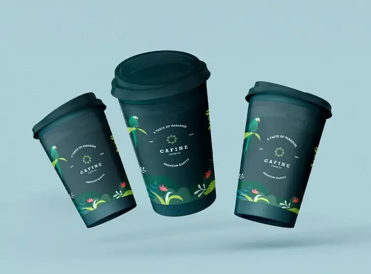 Three dark coffee cups with lids featuring a tropical design with a green parrot and colorful plants, labeled 'Cafine Coffee Co' and 'A Taste of Paradise Premium Quality.'
