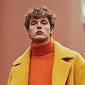 Young man with curly hair wearing an orange turtleneck and yellow coat against a blurred brown background.