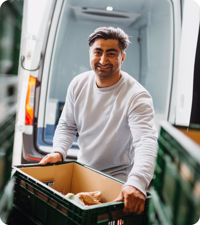 Smiling man in a grey sweatshirt loading a green crate with groceries into a van.