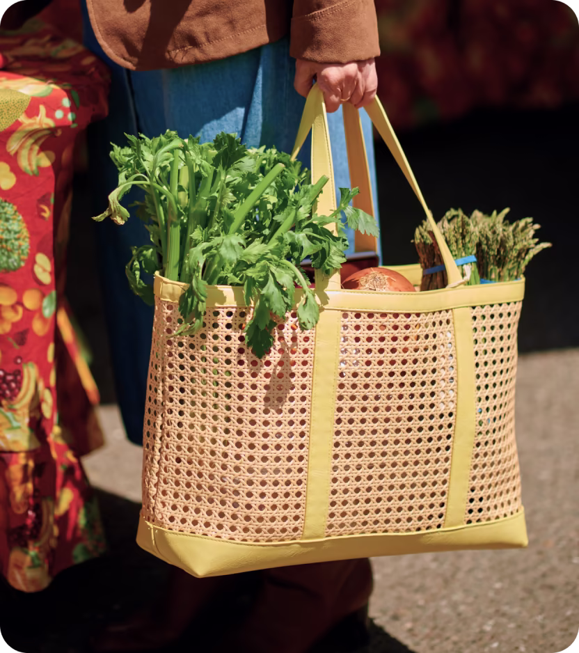Person holding a woven basket bag filled with fresh celery, onions, and bundles of asparagus.