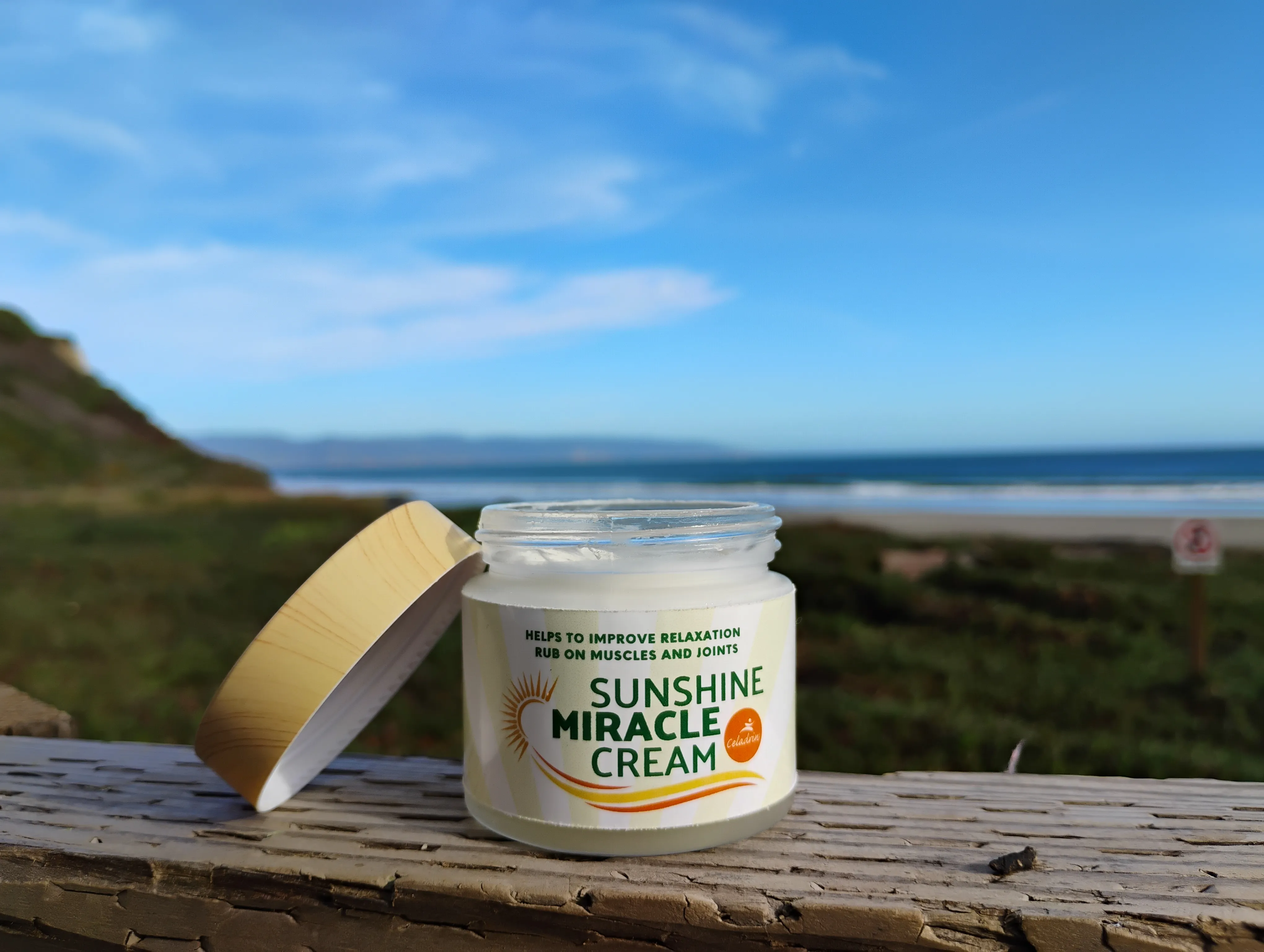Open jar of Sunshine Miracle Cream for muscle and joint relaxation on a wooden railing with a beach and blue sky in the background.