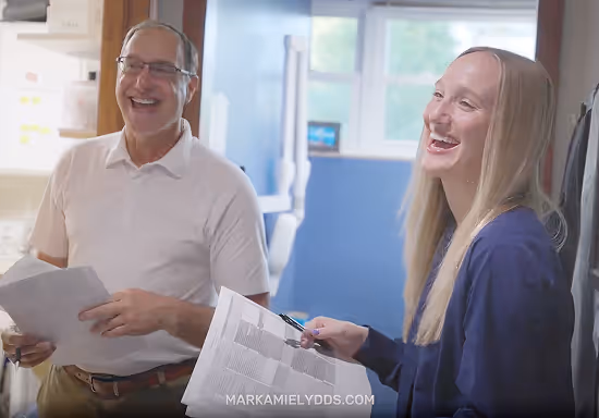Smiling man and woman holding papers together in a bright room, sharing a light moment.