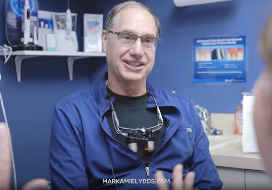 Smiling male dentist wearing glasses and dental loupes in a dental office with dental care products and posters in the background.