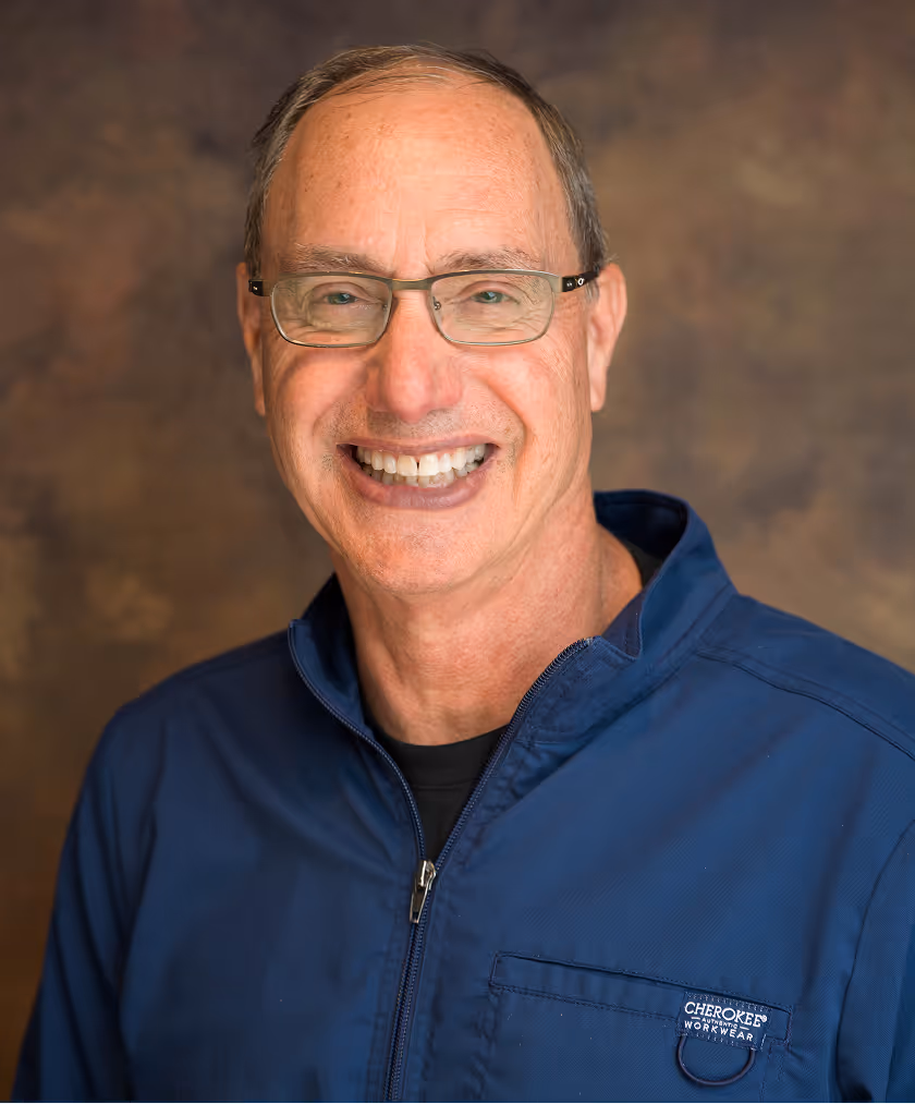 Smiling middle-aged man wearing glasses and a white polo shirt standing outdoors with a garden and wooden fence in the background.