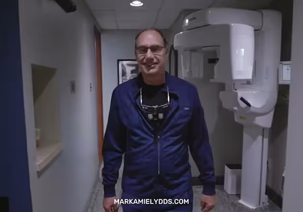 Smiling male dentist in navy scrubs standing next to dental imaging equipment in a clinic hallway.