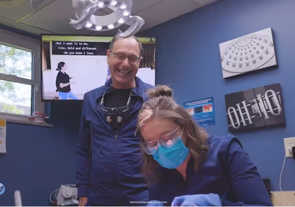 Smiling male dentist standing next to a female dental assistant wearing a mask and gloves in a dental office.