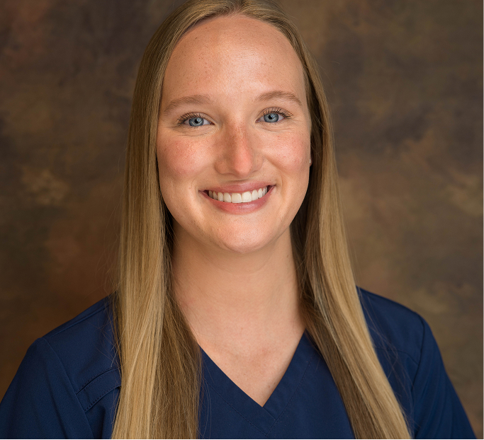 Smiling woman with long blonde hair wearing a blue scrub top against a brown backdrop.