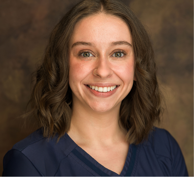 Smiling woman with shoulder-length curly brown hair wearing a navy blue top against a brown background.