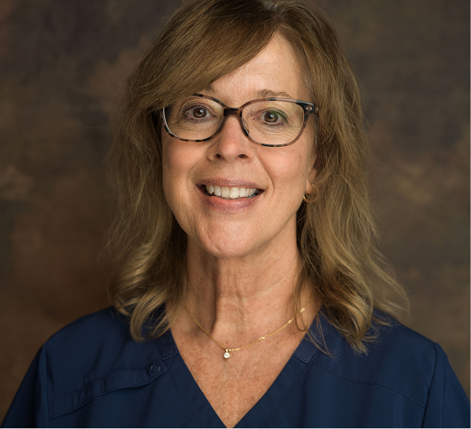 Smiling middle-aged woman with glasses and wavy brown hair wearing a navy blue top and a gold necklace.