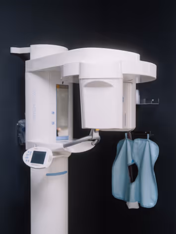 CT scanner machine with control panel and attached blue protective aprons against a dark wall.