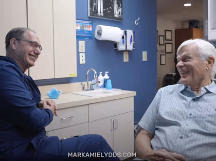 A dentist and an elderly male patient smiling and having a friendly conversation in a dental office.
