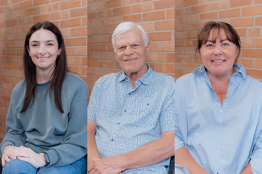 Portraits of three smiling people sitting against a brick wall background: a woman with long dark hair in a green sweatshirt, an older man with white hair in a light blue patterned shirt, and a woman with brown hair in a light blue button-up shirt.