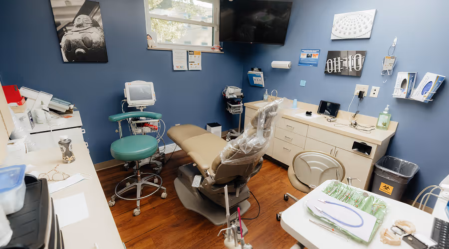 Dental exam room with a beige dental chair, green stool, dental tools on a tray, and blue walls decorated with framed pictures.