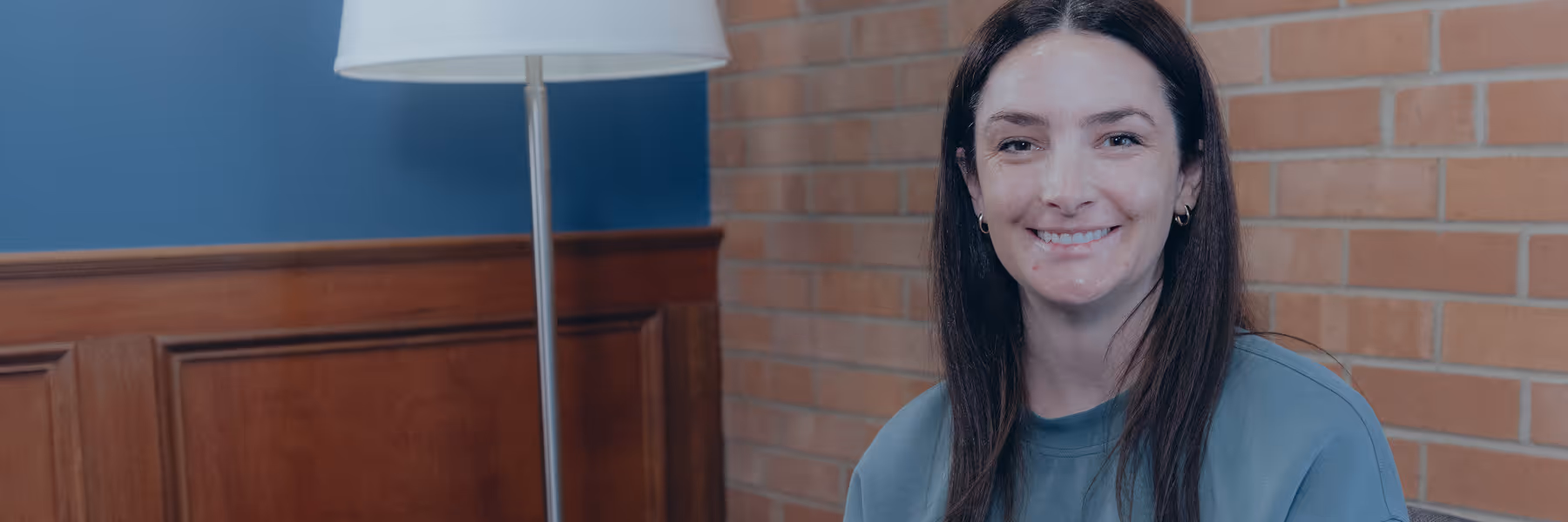 Smiling woman with long brown hair sitting indoors against a brick wall and wooden paneling with a floor lamp nearby.