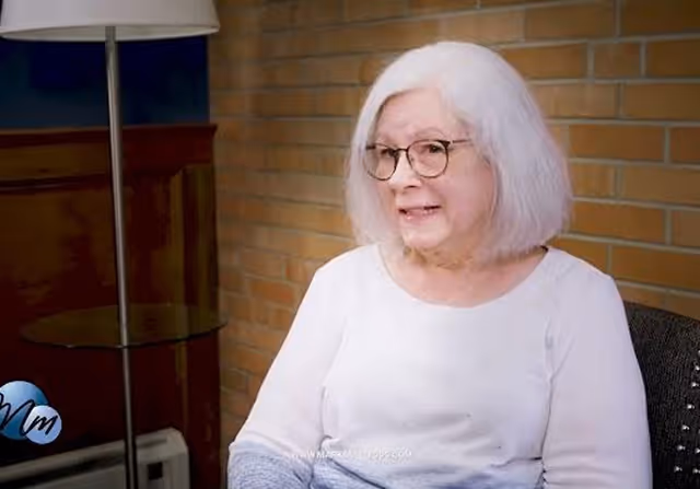 Elderly woman with white hair and glasses sitting in a chair against a brick wall, smiling.