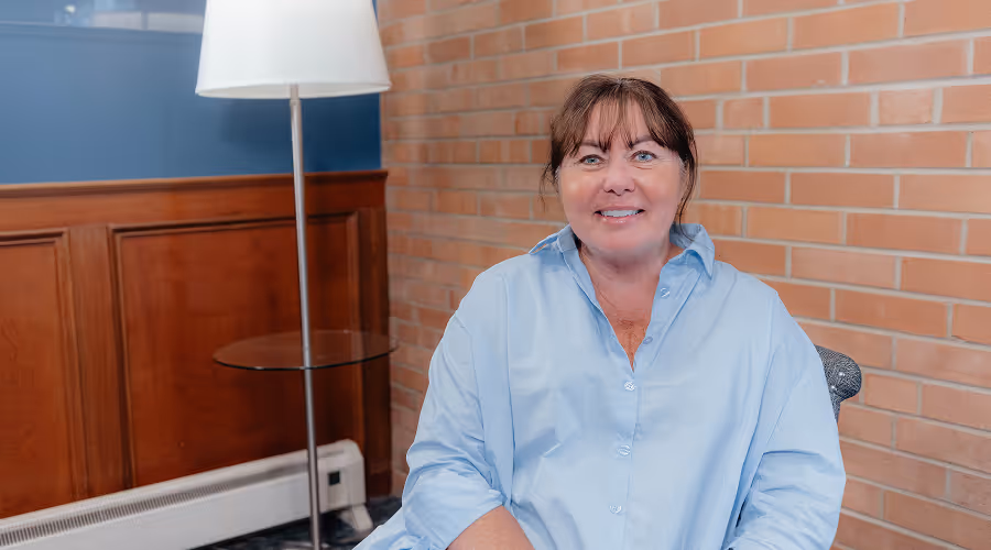 Smiling middle-aged woman with brown hair wearing a light blue shirt sitting against a brick and wood-paneled wall with a floor lamp nearby.