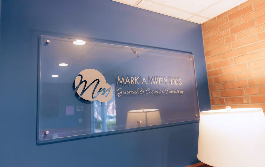 Transparent wall sign with logo and text reading 'Mark A. Miely DDS General & Cosmetic Dentistry' against a blue wall with a lamp and brick wall nearby.