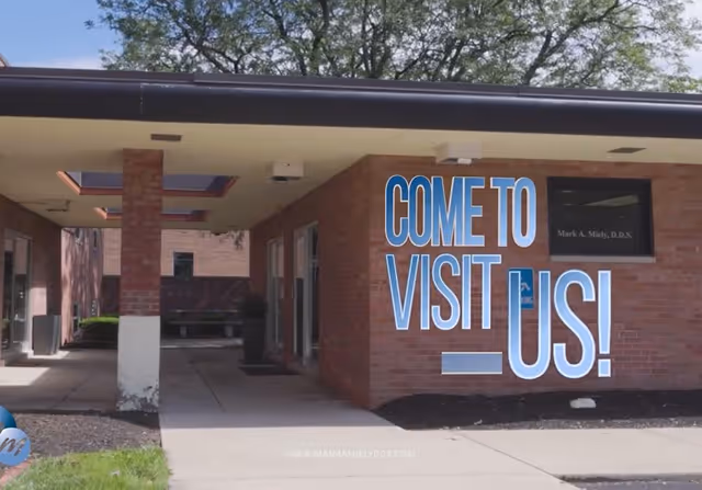 Entrance of a brick building with a covered walkway and a sign reading 'COME TO VISIT US!' in blue letters on the wall.