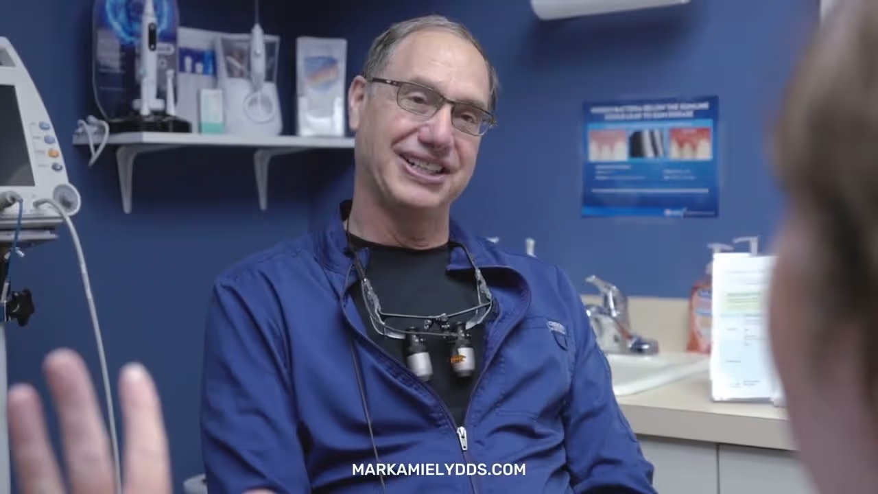 Smiling male dentist wearing glasses and a blue jacket with dental loupes around his neck, sitting in a dental office.