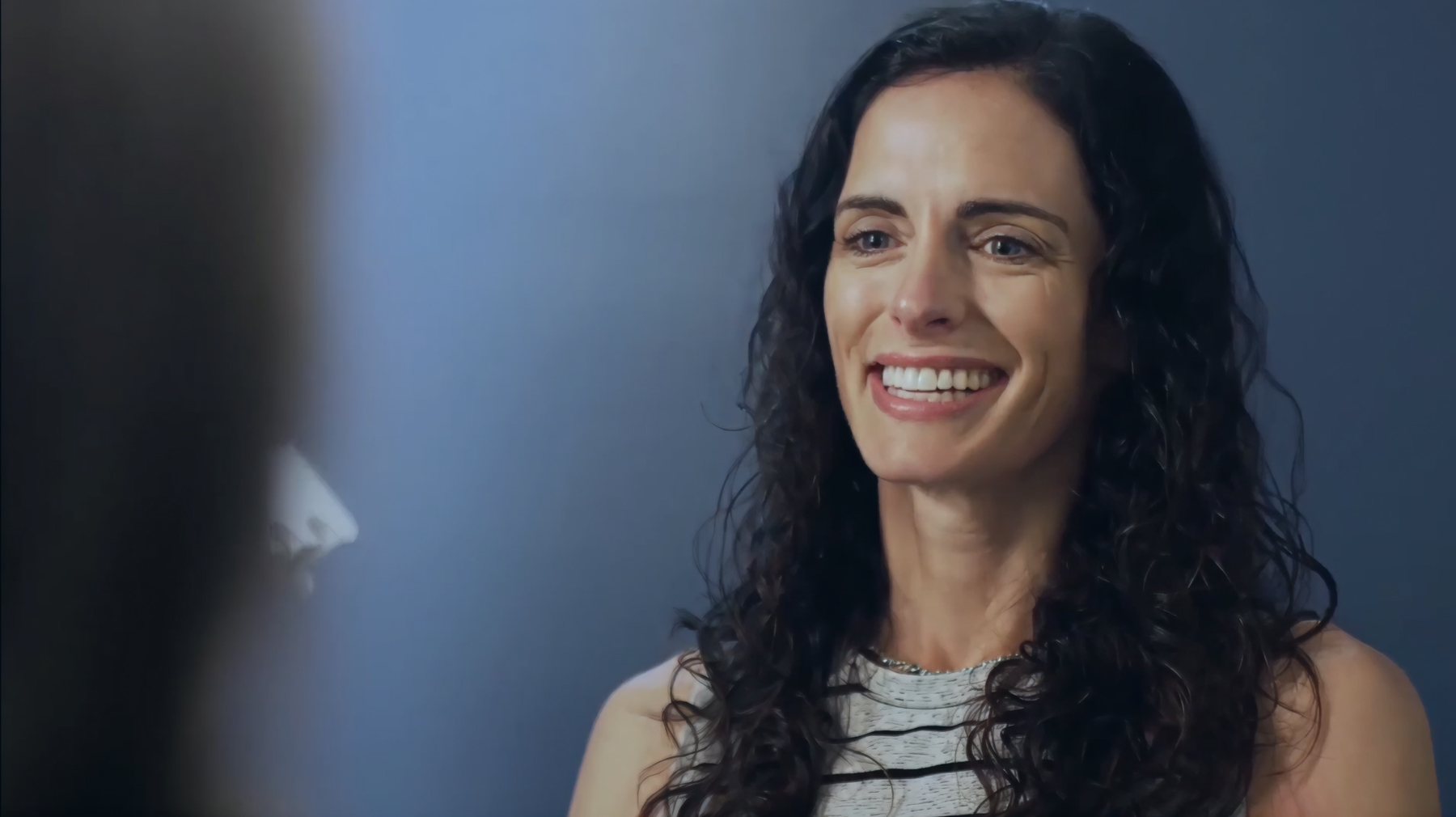 Smiling woman with long curly hair wearing a striped sleeveless top against a blue background.