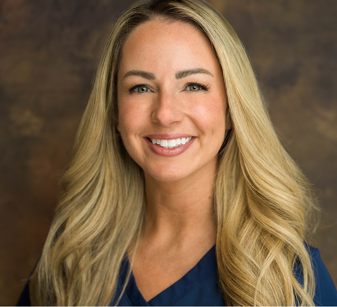 Portrait of a smiling woman with long blonde hair wearing a dark blue top against a brown blurred background.