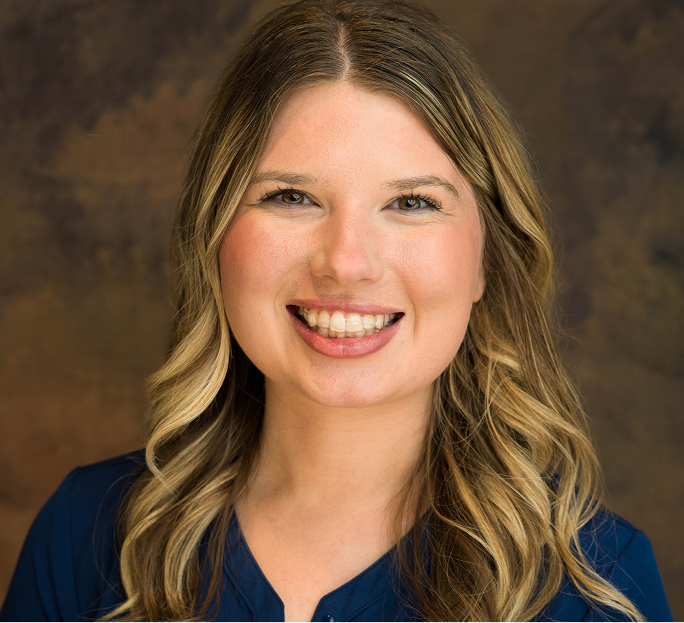 Portrait of a smiling woman with long wavy blonde hair wearing a dark blue top against a brown background.