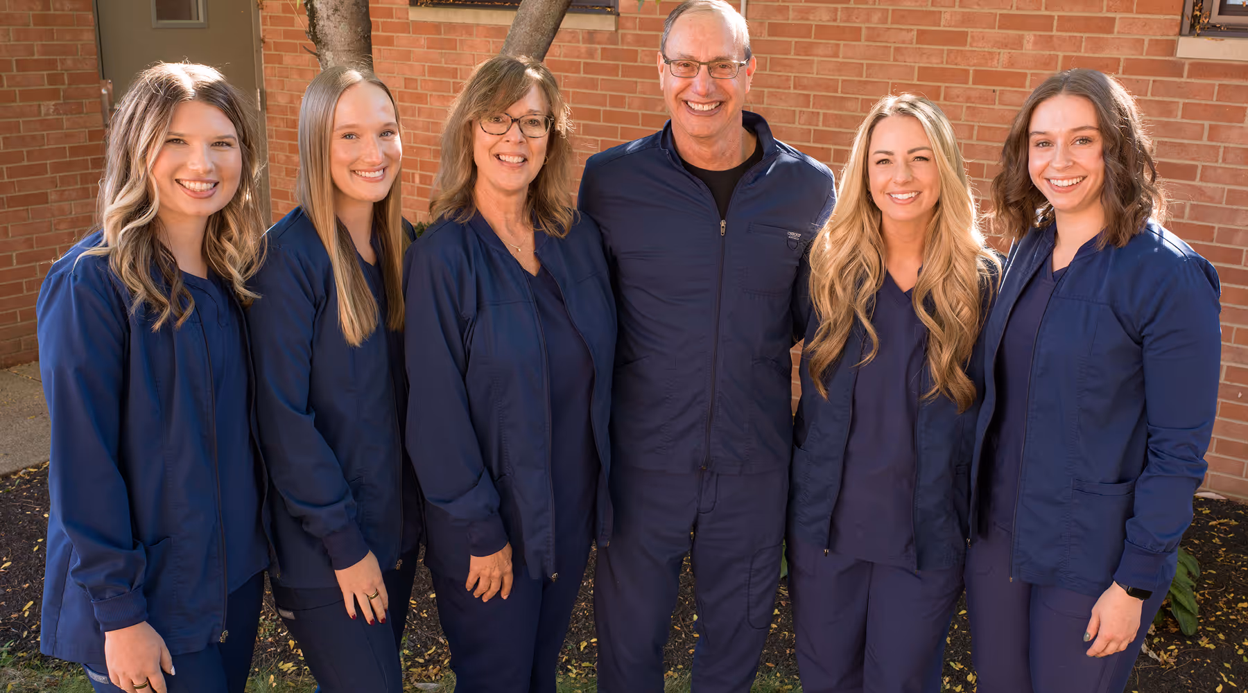 Group of six smiling healthcare professionals wearing navy blue scrubs standing outdoors in front of a brick wall.