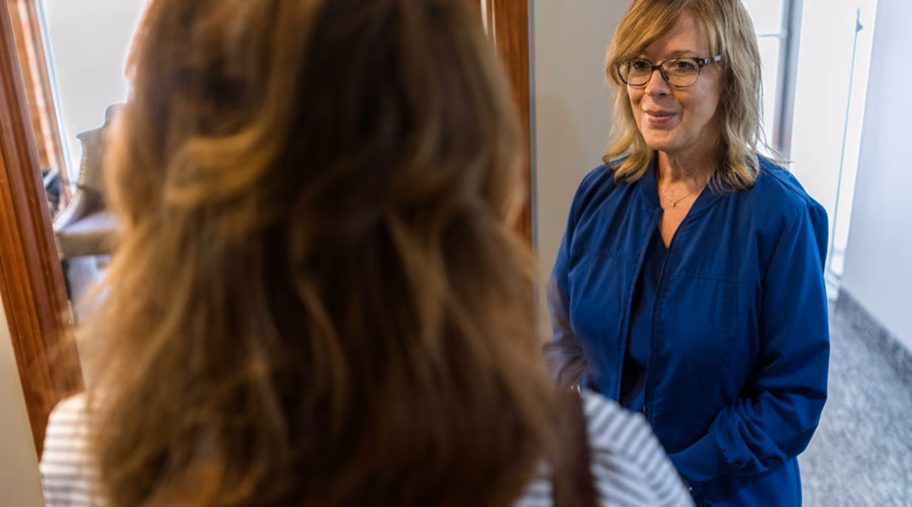 A female healthcare professional in blue scrubs smiling and talking with a woman seen from behind.