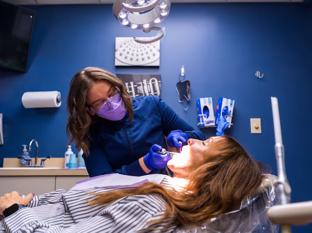 Dentist wearing purple mask and gloves examining a patient's mouth in a dental clinic with blue walls.