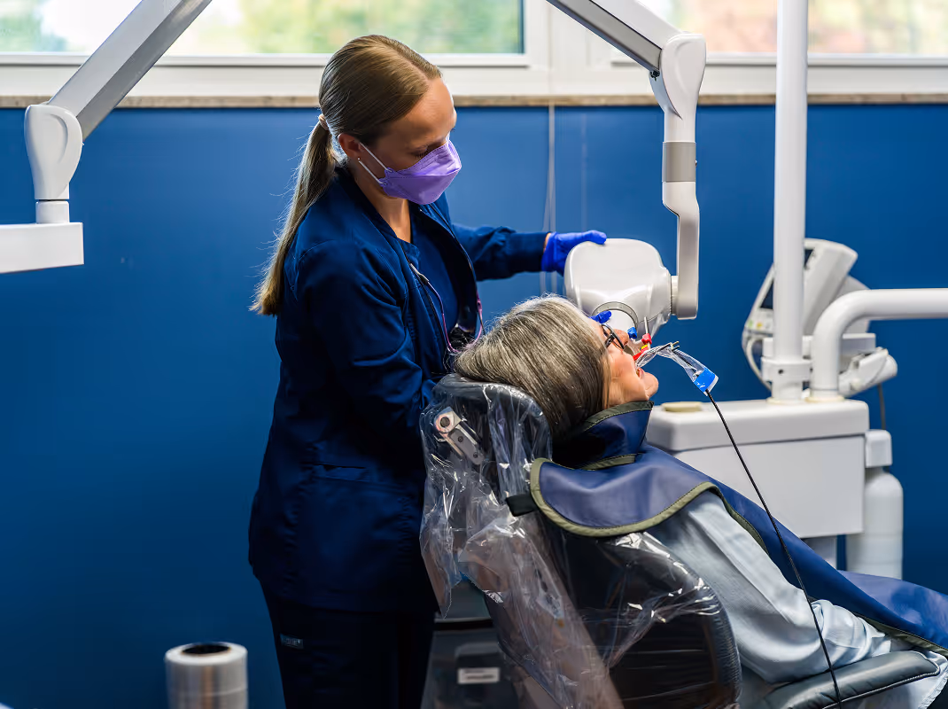 Dental professional wearing a purple face mask positioning an X-ray machine over a patient seated in a dental chair.