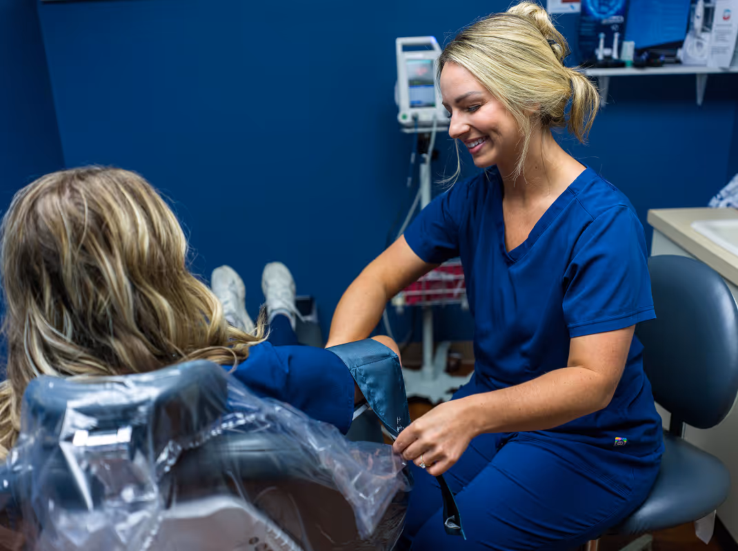 Healthcare professional in blue scrubs smiling while putting a blood pressure cuff on a patient seated in a chair.