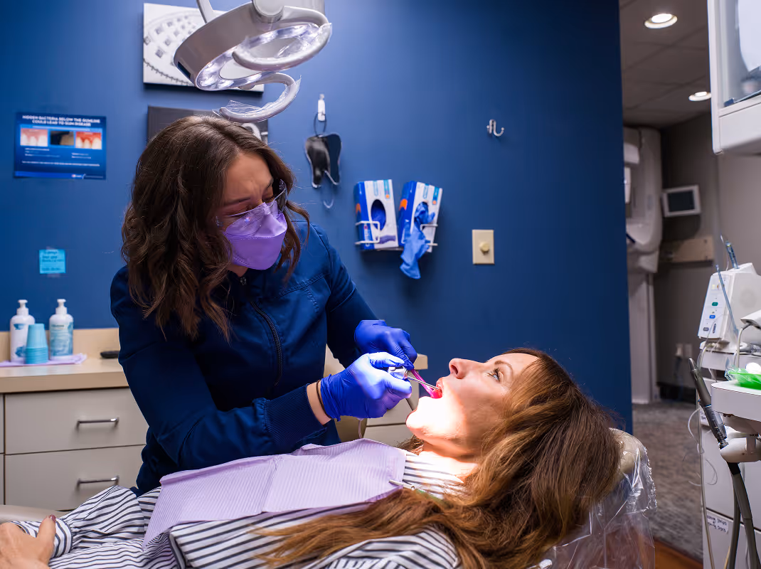 Dentist wearing purple mask and gloves performing dental treatment on a female patient lying in a dental chair.
