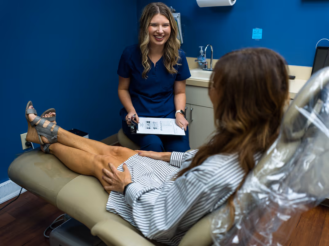 A smiling healthcare professional in navy scrubs holds a clipboard and talks with a seated patient in a medical office.