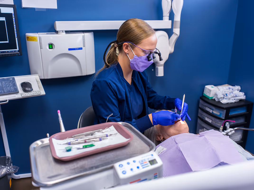 Dentist wearing purple mask and magnifying glasses treats patient reclining in dental chair in clinic.
