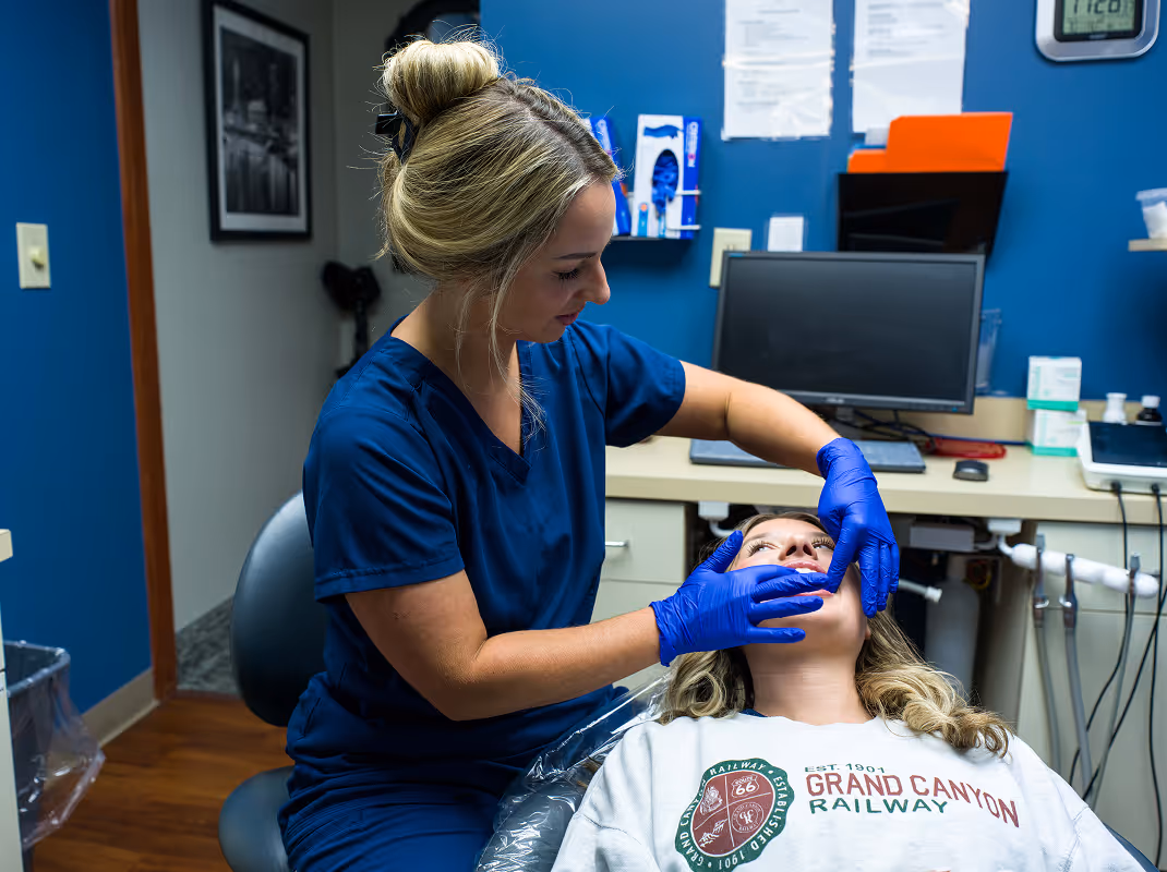 Dental professional wearing blue scrubs and gloves examining a patient's teeth in a dental office.