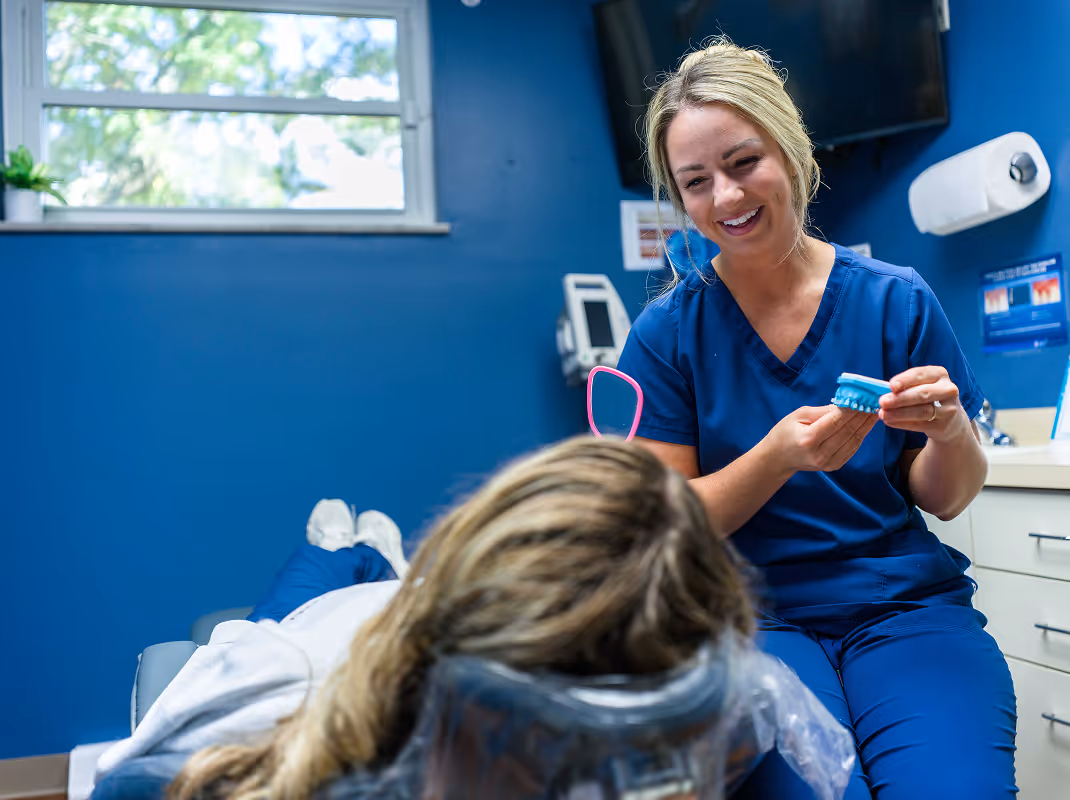 Female dental professional in blue scrubs smiling and explaining a dental mold to a patient lying back in a dental chair.