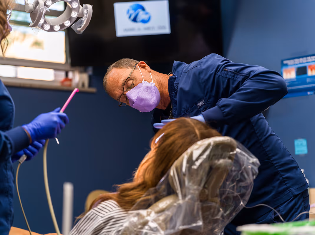 Dentist wearing a purple mask and glasses examining a patient's mouth while an assistant holds dental tools in a blue-walled clinic.