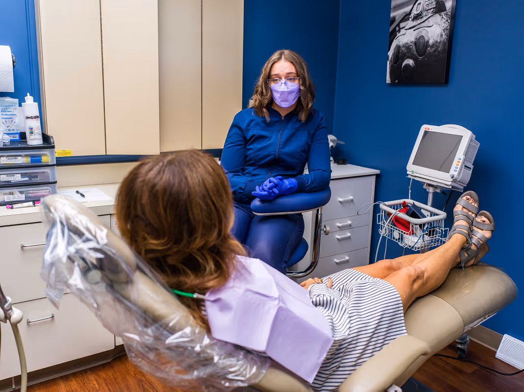 Dentist wearing blue scrubs, gloves, and a purple mask talks to a patient reclining in a dental chair in a clinic room.