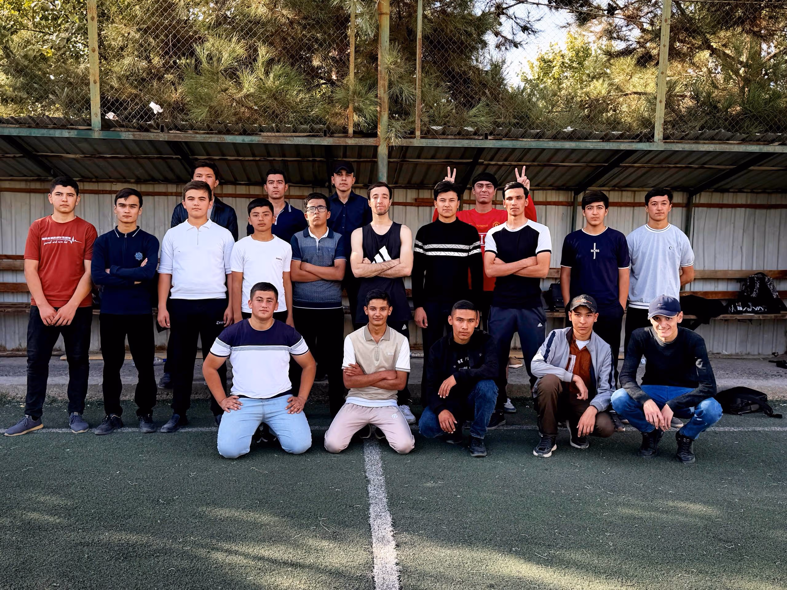 Group of young men posing on a sports field with a metal shelter and trees in the background.