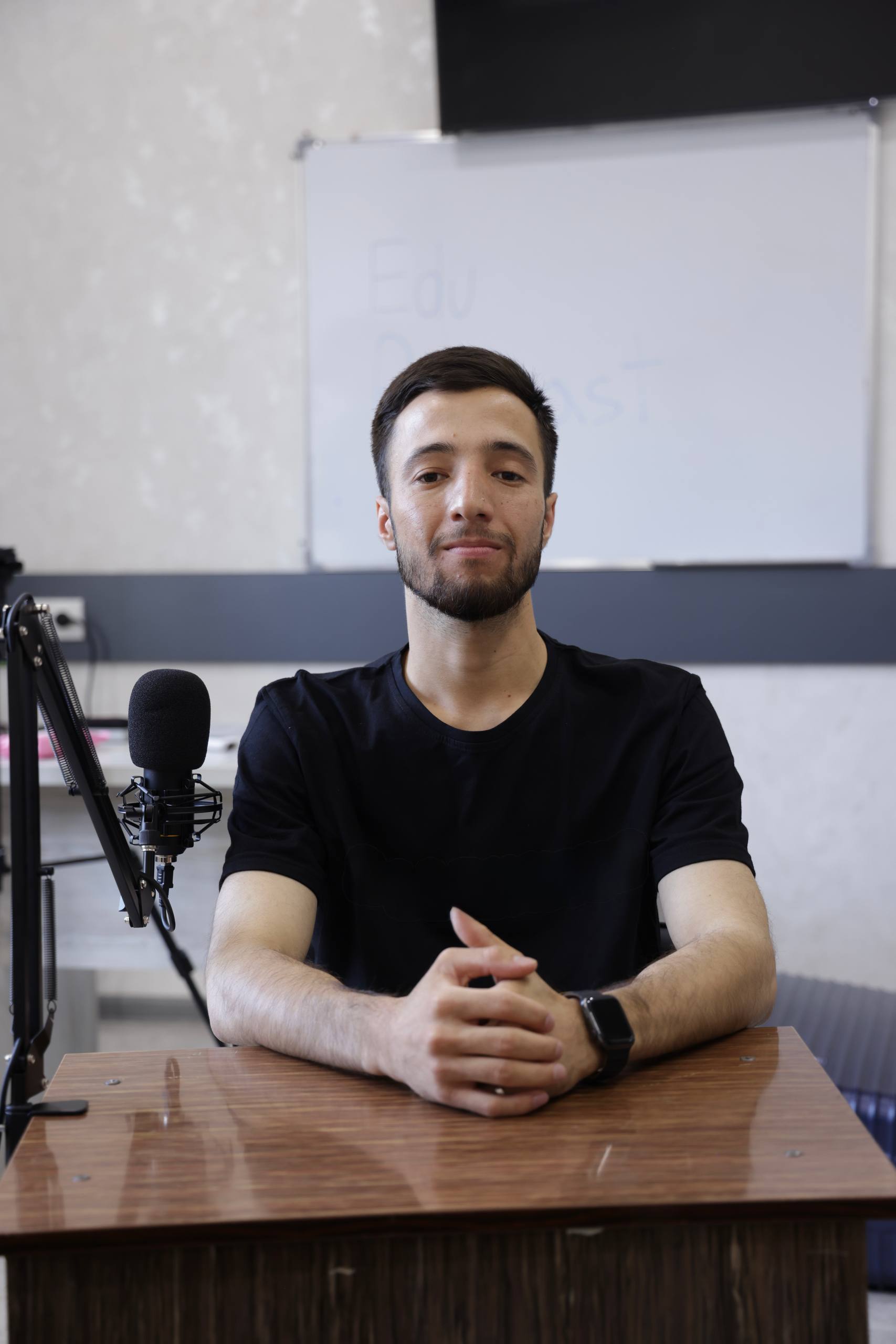Man with beard sitting at a wooden desk with a microphone, facing the camera.