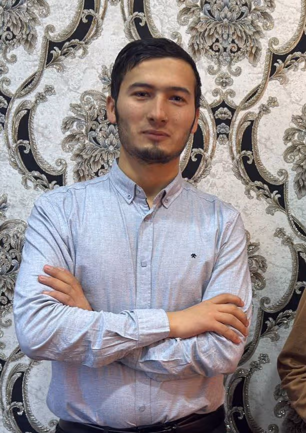 Young man with short dark hair and a trimmed beard wearing a light gray button-up shirt standing with arms crossed in front of an ornate floral patterned wallpaper.