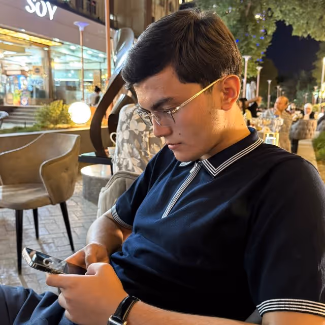Young man wearing glasses and a black polo shirt sitting outside at night, focused on his smartphone.