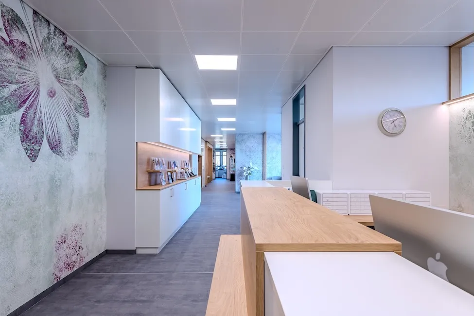 Moderner Praxisflur des Praxisstandort in Kaufbeuren mit heller Beleuchtung, white Cabinets und einem großen Holztresen, Wand mit Blumendekor links.