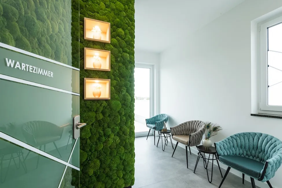Heller Warteraum in der dermatologische Praxis in Landsberg am Lech mit grüner Mooswand, drei eingelassenen Vasen mit Licht und modernen gepolsterten Stühlen.