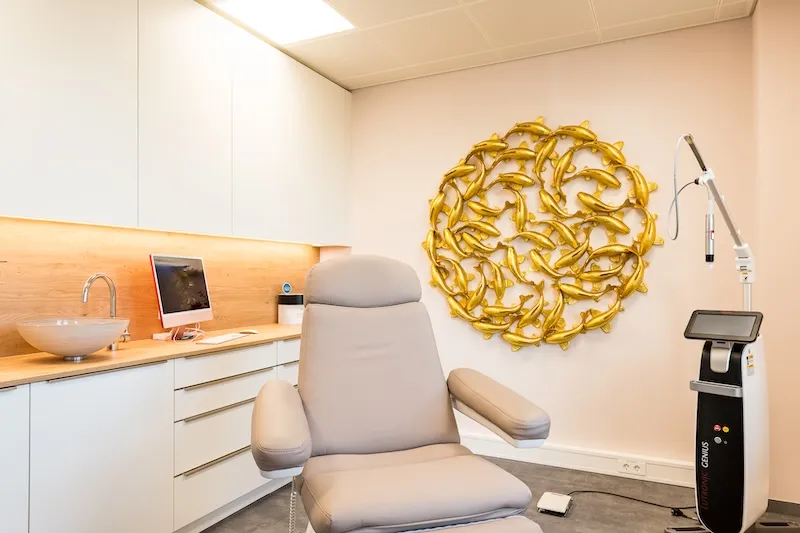 Ein moderner Behandlungsraum des Praxisstandort Kaufbeuren mit einem grauen Behandlungsstuhl, einem Computermonitor auf einer Holzarbeitsplatte und einer goldfarbenen Wanddekoration aus Fischen.